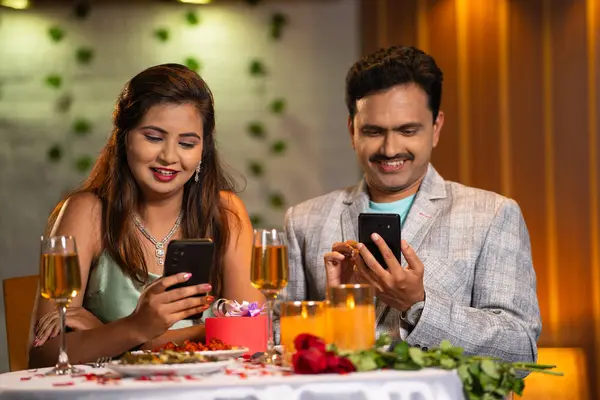 Indian couples at romantic candlelight dinner busy using mobile phone at restaurant - concept of social media sharing and technology distraction.