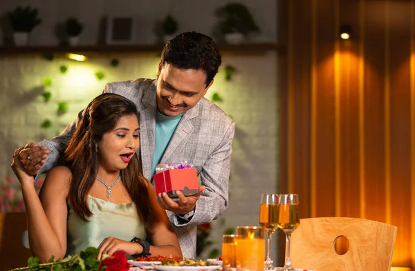 happy husband giving surprise gift to wife at candlelight dinner for anniversary or birthday - concept of romantic date night, relationship bonding and affection.