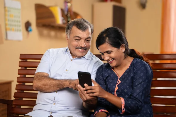 50s Indian father showing proposal photos on mobile phone to daughter for marriage at home - concept of match making, arranged marriage and responsibility.