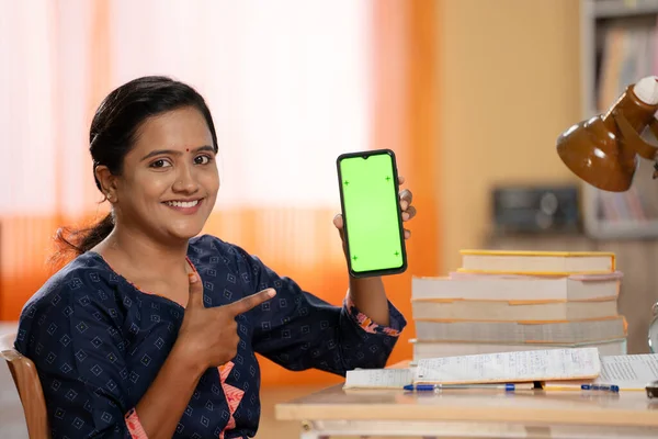 Indian young girl showing green screen mobile phone while reading at study table at home - concept of online education, e learning and virtual learning.