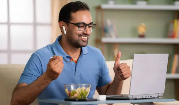 Happy indian man eating fruit salad by showing thumbs up on online video call at home - concept of satisfaction, communication and healthy eating