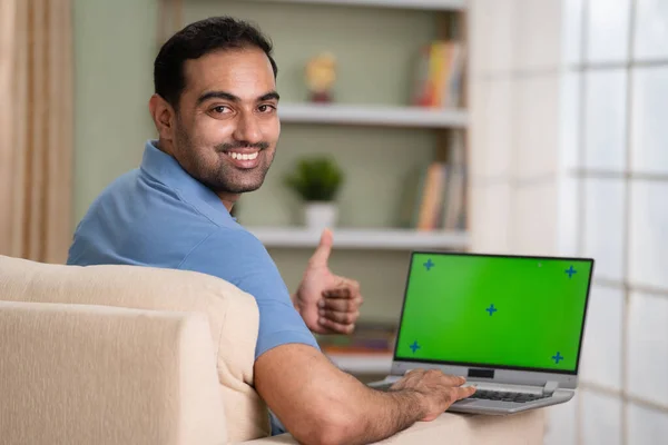 Happy indian man using green screen laptop by showing thumbs up while looking at camera at home - concept of satisfaction, approval and advertisement.