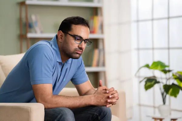 Worried indian man deep thinking while sitting on sofa at home - concept of financial problem, love breakup and mental illness