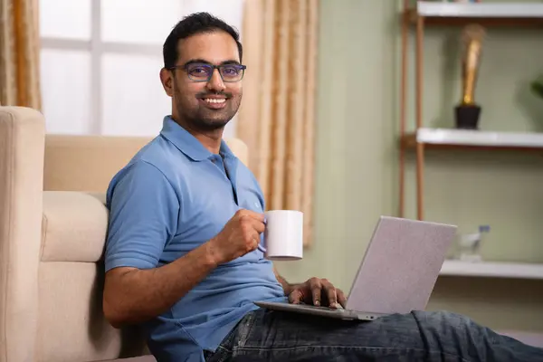 Happy indian man using laptop by drinking coffee or tea by looking at camera while sitting on floor at home - concept of refreshment, technology and weekend holidays.