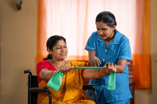 Indian nurse or therapist guiding or assisting Woman on wheelchair to stretching elastic band at home - concept of health care service, professional occupation and physiotherapist