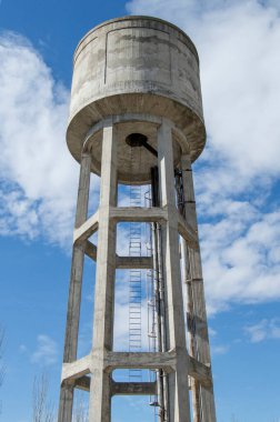 Aşağıdan su tankı görünüyor ve bulutlu mavi gökyüzüne karşı siluetlenmiş.