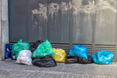 Metal bir kapının yanında birkaç renkli çöp torbası var.