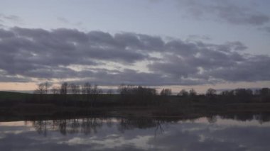 Evening by the lake in the countryside. A cloudy twilight sky is reflected on the surface of the lake water. The bare silhouettes of the trees suggest the late fall season.