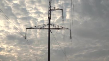 Yüksek voltajlı direğin karanlık siluetleri kırsal panoramik bir arazide güneşli, bulutlu bir gökyüzü arka planında görülüyor..