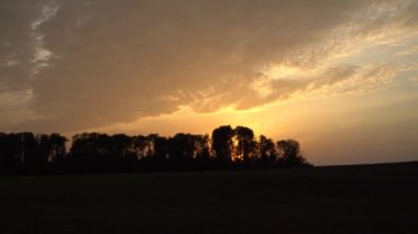 Karanlık ağaç siluetlerinin arkasında, panoramik bir manzarada, alevli günbatımı ışığı. Bu batan cennet manzarasının üzerinde dramatik bulutlu bir gökyüzü var. Alacakaranlık saatinden önce..