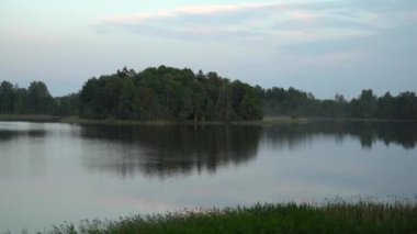 Sakin bir gölde, sulara yansıyan yemyeşil bir adayla huzurlu bir yaz akşamı. Gökyüzü hafif bulutlu, sakin manzaraya huzur katıyor..
