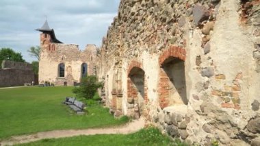 Letonya 'daki Dobele Kalesi harabeleri, tarihi taş duvarlar, kemerli pencereler, ortaçağ kulesi ve yazın parlak gökyüzünün altında bankları olan yemyeşil bir çimenlik..