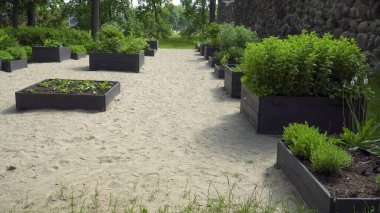 Çeşitli bitki ve otlarla dolu birden fazla yükseltilmiş yatakları olan bir halk bahçesi. Bahçe, ağaçlar ve taş duvarlarla çevrili kumlu bir alana kurulmuş, sakin ve düzenli bir ortam yaratıyor..