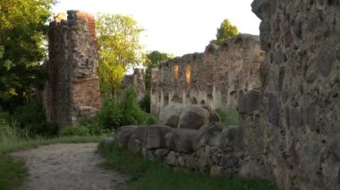 Güneş ışığıyla yıkanmış eski taş kalıntılarının sakin manzarası, aşırı gelişmiş bitki örtüsü ve kırsal bir toprak yol boyunca uzanan büyük kayalar, tarih ve doğa duygusunu çağrıştırıyor..