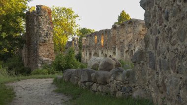 A serene view of ancient stone ruins with crumbling walls bathed in sunlight, surrounded by overgrown vegetation and large boulders along a rustic dirt path, evoking a sense of history and nature. clipart