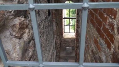 Eski tuğla ve taş duvarlı dar bir ortaçağ geçidi, metal parmaklıklarla çevrili, dışarıya bakan bir pencereye açılıyor. Tarihi atmosfer derinlik ve entrika ekler..