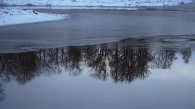 Sükûnetli bir kış sahnesinde kısmen donmuş bir nehir, açık bir şekilde karanlık bir gökyüzüne karşı çıplak ağaçların yansımaları görülüyor. Karla kaplı kıyılar ve ince buz kalıpları sakin atmosfere katkı sağlar..