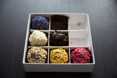 Her biri fındık, hindistan cevizi talaşı, çikolata ve çiçek yaprakları gibi malzemelerle süslenmiş çeşitli trüf mantarları içeren beyaz bir kutu. Koyu ahşap bir masa arkaplanına karşı ayarla.