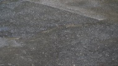 Yağmur damlaları asfalt bir yüzeye vurarak küçük su akıntıları ve su birikintileri oluşturur. Asfaltın ıslak dokusu ve dağınık damlacıklar yağmurlu havayı vurgular..