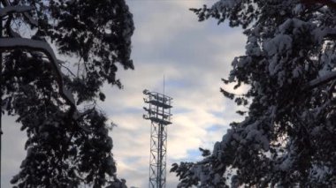5G mobil iletişim kulesi karla kaplı çam dalları arasında duruyor. Bir kış akşamı bulutlu bir günbatımı gökyüzü ile çekilen sahne, teknoloji ve doğa arasındaki zıtlığı vurgular..