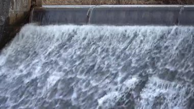 Şiddetli bir su akıntısı barajın üzerinde çağlayarak türbülans deseni oluşturur. Barajın beton yapısı belirgin bir şekilde, insan mühendisliği ve doğal güçler arasındaki etkileşimi vurguluyor..