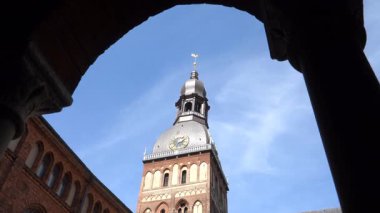 Tarihi Riga Dome Kilisesi kulesi, ikonik saati ve rüzgar gülüyle mavi gökyüzüne doğru yükseliyor. Kavisli bir geçitten alçak açılı bir açıdan görüntülendi..