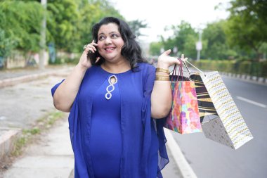 Genç Hintli, elinde alışveriş torbalarıyla sokakta yürüyen iri yarı bir kadın. Şişman kadın telefonda konuşuyor ve alışveriş yapıyor..