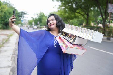 Genç Hintli artı boyunda bir kadın alışveriş poşetleri tutuyor. Açık havada akıllı telefonuyla selfie çekiyor. Telefonu kullanan şişman kadın..