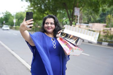 Genç Hintli artı boyunda bir kadın alışveriş poşetleri tutuyor. Açık havada akıllı telefonuyla selfie çekiyor. Telefonu kullanan şişman kadın..