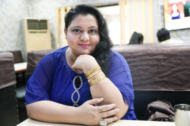 Genç ve cüsseli Hintli bir kadın restoranda oturmuş yüzünde gülümsemeyle kameraya bakıyor..