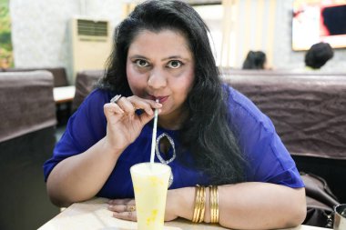 Genç ve cüsseli Hintli bir kadın restoranda oturmuş smoothie içiyor..