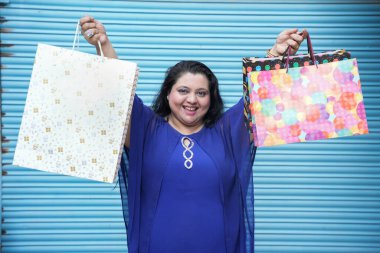 Mavi arka planda izole edilmiş alışveriş torbaları taşıyan genç, neşeli Hintli, iri yarı bir kadın. Kilolu kadın alışverişi. konsept.