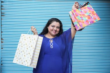 Mavi arka planda izole edilmiş alışveriş torbaları taşıyan genç, neşeli Hintli, iri yarı bir kadın. Kilolu kadın alışverişi. konsept.