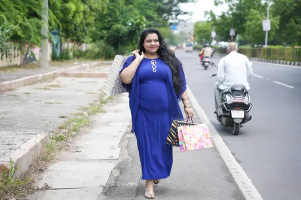 Genç Hintli, elinde alışveriş torbalarıyla sokakta yürüyen iri yarı bir kadın. Şişman kadın alışveriş yapıyor..