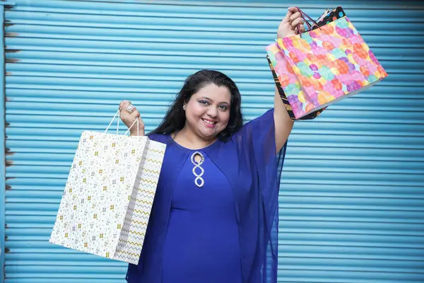 Mavi arka planda izole edilmiş alışveriş torbaları taşıyan genç, neşeli Hintli, iri yarı bir kadın. Kilolu kadın alışverişi. konsept.