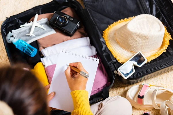 旅行のためにパックするもののチェックリストを作る 女性の執筆紙はノートを取り 休暇にスーツケースを梱包する部屋に座っている紙のリストを書く 荷物に服を準備する 旅行休暇旅行 — ストック写真
