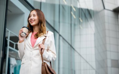 Happy business woman smiling hold paper cup of hot drink outdoor walking on street, Asian businesswoman holding coffee cup takeaway going to work she walking near her office building outside