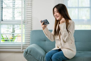 Heyecanlı kadın cep telefonuyla yumruk kaldırıyor zaferi kutluyor online kazanıyor ya da evdeki kanepede iyi haberler alıyor mutlu Asyalı kadın akıllı telefonda komik ve heyecanlı.