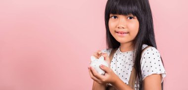 Asyalı küçük bir çocuk kumbarasını tutuyor ve pembe arka planda izole edilmiş stüdyo çekimlerine bakıyor, mutlu bir çocuk gibi gülümsüyor, tam bir domuz kumbarası, kişisel tasarruf