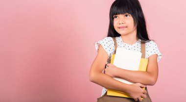 Asyalı küçük bir çocuk, 10 yaşında. Stüdyoda okula gitmek için kitap tutuyor. Pembe arka planda izole edilmiş, mutlu kız çocuğu ders kitaplarına sarılıyor.