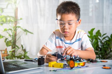 Asyalı çocuk, Arduino robot arabasıyla sensör çipine enerji ve sinyal kablosu takıyor, küçük çocuk oyuncaklarla uzaktan öğreniyor, STEAM eğitim teknolojisi dersleri.