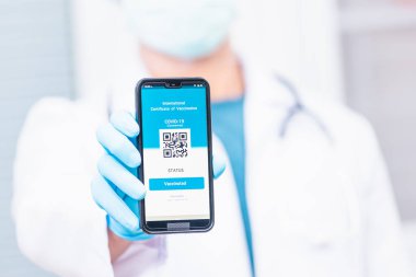 Tıbbi maskeli doktor, uygulamalı akıllı telefon dijital aşılı pasaport Coronavirus COVID-19 sertifikası gösteriyor.