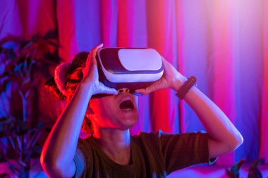Asyalı kadın VR başlığını takıyor. Evde heyecanlı duygusal bir şekilde mor ve mavi arka plan, sanal gerçeklik gözlüğü takan kadın gerçekliği, metazeni ve siber uzay kavramını deneyimliyor.
