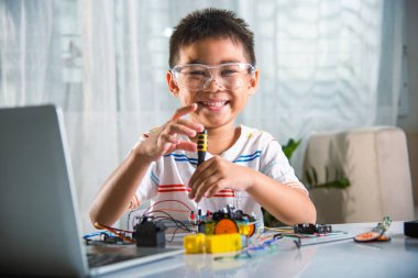 Asian kid boy assembling the Arduino robot car homework project at home, Little child tighten the nut with a screwdriver to assemble car toy, creating electronic AI technology workshop school lesson