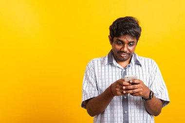 Asyalı mutlu bir portre genç siyah adam ayakta gülümsüyor cep telefonuyla sosyal mesaj yazarken veya okurken cep telefonu boş ekranında, stüdyo sarı arka planda fotokopi alanı ile çekim yapıyor.