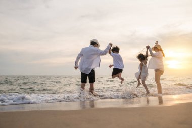 Yolculuğun ve tatilin tadını çıkaran insanlar aile babası, annesi, oğlu ve kızının silueti plajda el ele tutuşurken mutlu aile gün batımında sahilde zıplayarak eğleniyor.