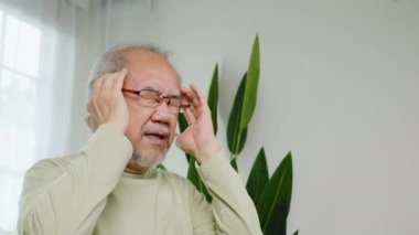 Baş ağrısı. Üzgün Asyalı son sınıf öğrencisi kronik migren ağrısı masajı, yaşlı adam baş ağrısı için elini tutuyor oturma odasında, yaşlılık sağlık sorunları, sağlık sorunları...