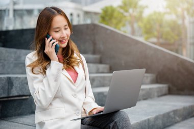 Genç Asyalı mutlu, serbest çalışan, dizüstü bilgisayarlı, gülümseyen, kahve fincanıyla cep telefonuyla konuşan, şehrin dışındaki merdivenlerde oturan genç bir kadın. Uzaktan çalışma konsepti ofis dışında.