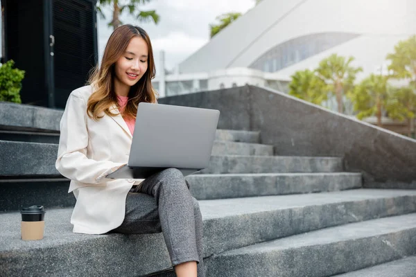Remote working concept outside the office. Happy young Asian woman freelance smile looking laptop computer with smile while working with coffee cup while sitting on stairs outdoors in the city.