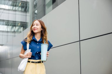 Asyalı güzel bir iş kadını elinde çelik termos bardak bardak tutan güler yüzlü modern bina ofisinin yanındaki sokakta yürüyen mutlu bir kadın.